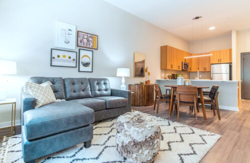 A living room with high ceilings and hardwood style flooring, leading into a kitchen with a breakfast bar and pendant lighting.